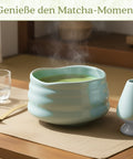 Gewurzladen Nafels Dampfender heißer Matcha Tee in einer hellblauen Schale neben einem Besenhalter und einem Löffel.